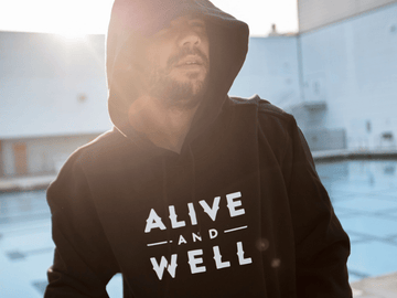 Person in a black hoodie with 'Alive and Well' logo, standing outdoors in sunlight.