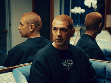 Person with a shaved head wearing a black 'Alive and Well' sweatshirt, sitting in front of mirrors.